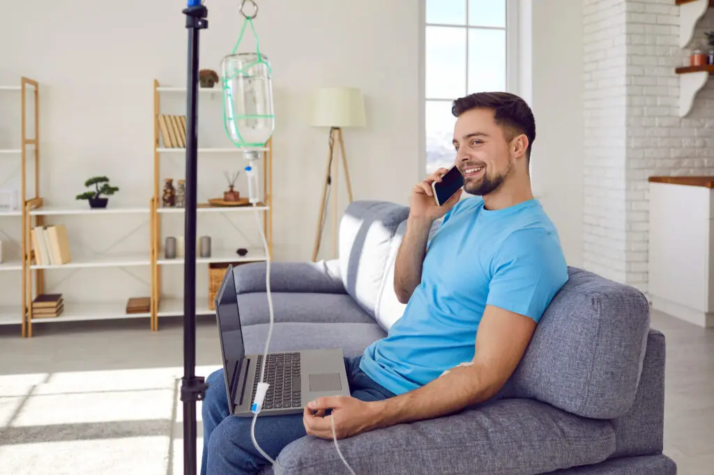 Young man talking by mobile phone sitting on sofa at home while receiving IV vitamin therapy.